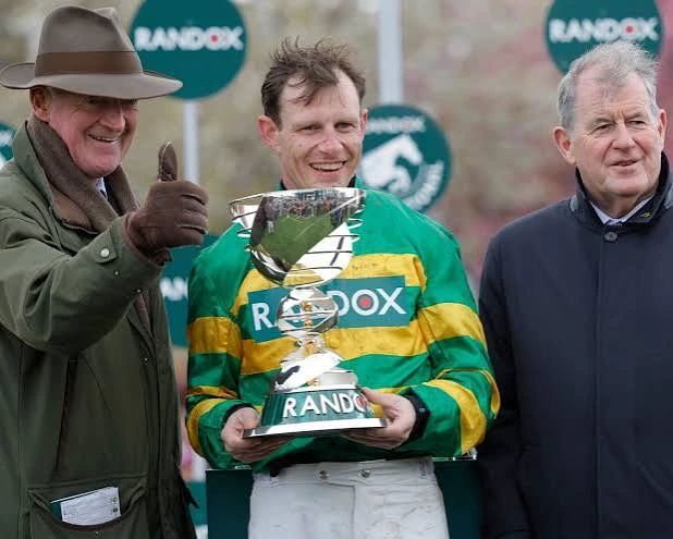 Jockey in green and gold silks holds a Randox trophy between two officials.