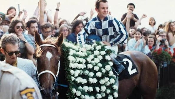 Jockey in checkered silks rides a flower-draped racehorse through a cheering crowd.
