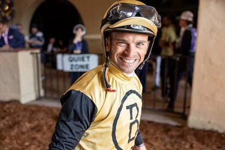 Smiling jockey in gold and black racing silks stands inside a stable.