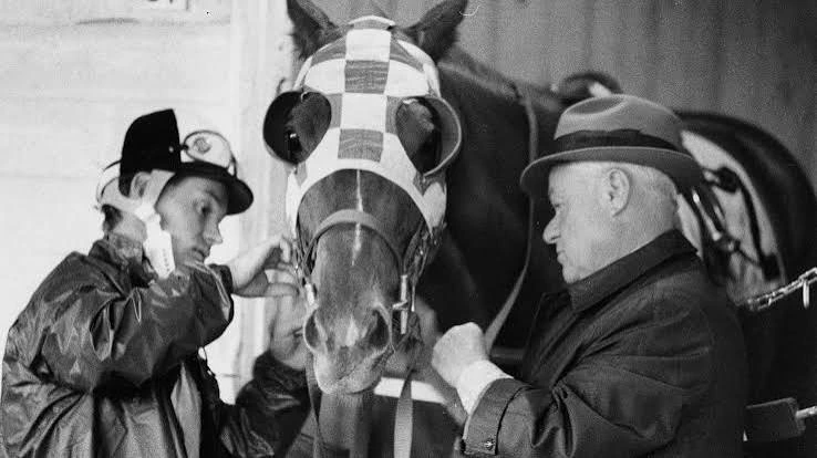 Jockey and trainer prepare a racehorse wearing checkered blinkers before competition.