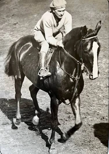 Jockey rides a dark racehorse with a white blaze in a historic photograph.